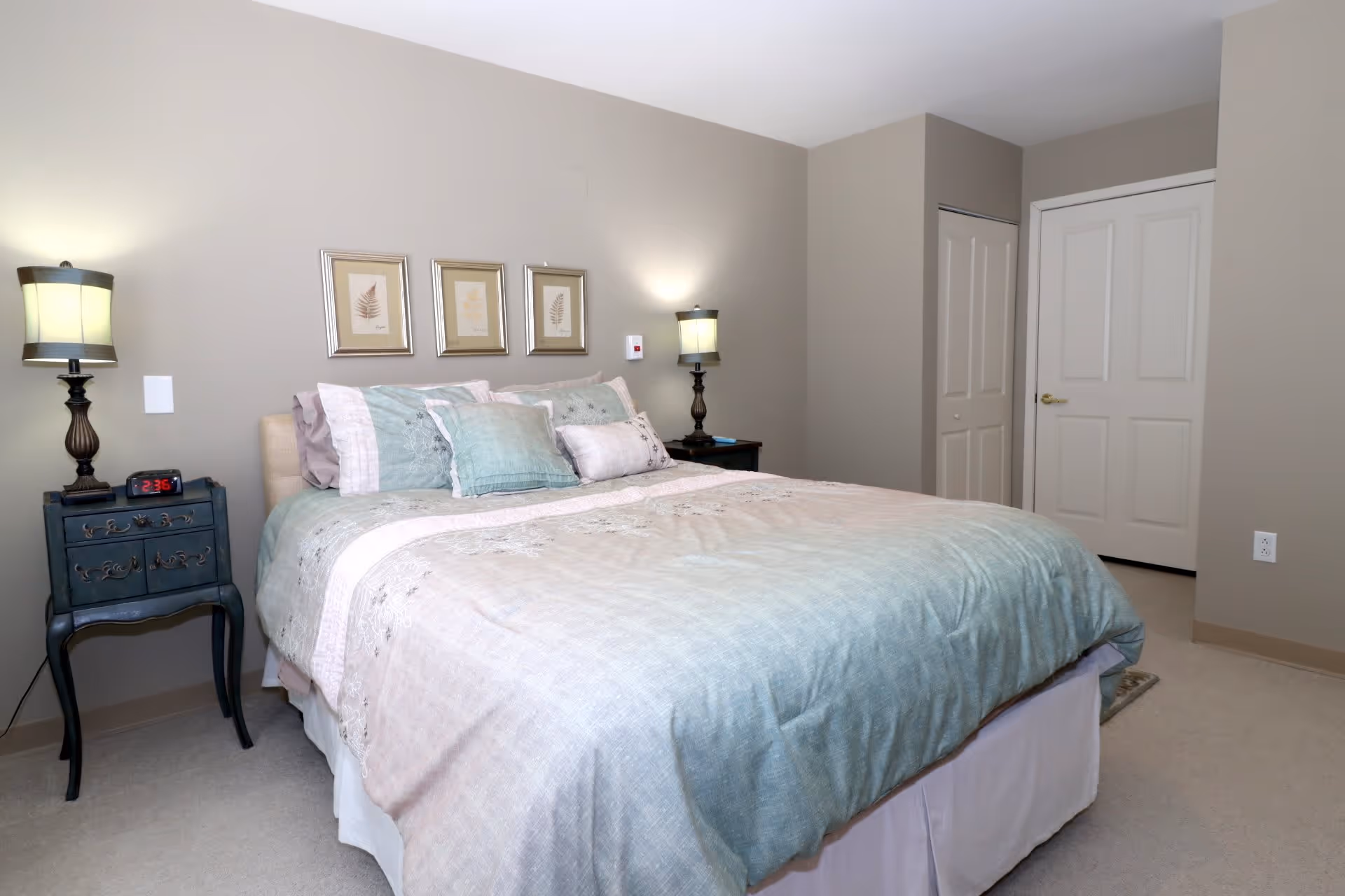 A neatly made bed with light blue and beige bedding in a bedroom. There are two bedside tables with matching lamps on each side of the bed. Three framed botanical prints hang on the wall above the bed. The room has beige walls and carpeted floor, with a closed closet door and a regular door visible in the background.