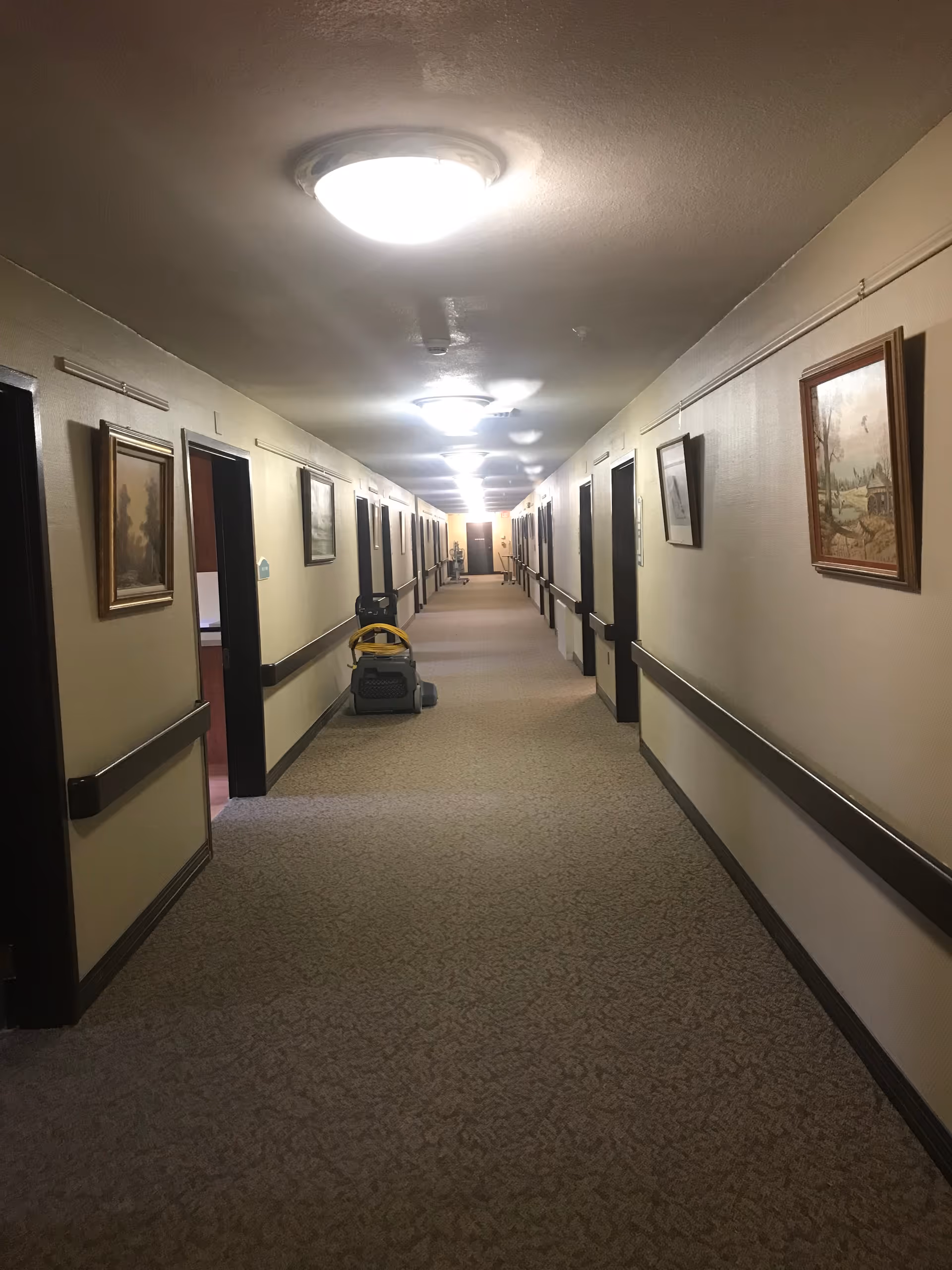 Long indoor hallway in a senior living facility with beige walls and carpeted floor. Several closed doors line both sides of the hallway, with handrails along the walls. Ceiling lights illuminate the corridor, and framed paintings hang on the walls. A cleaning machine is parked on the left side of the hallway.
