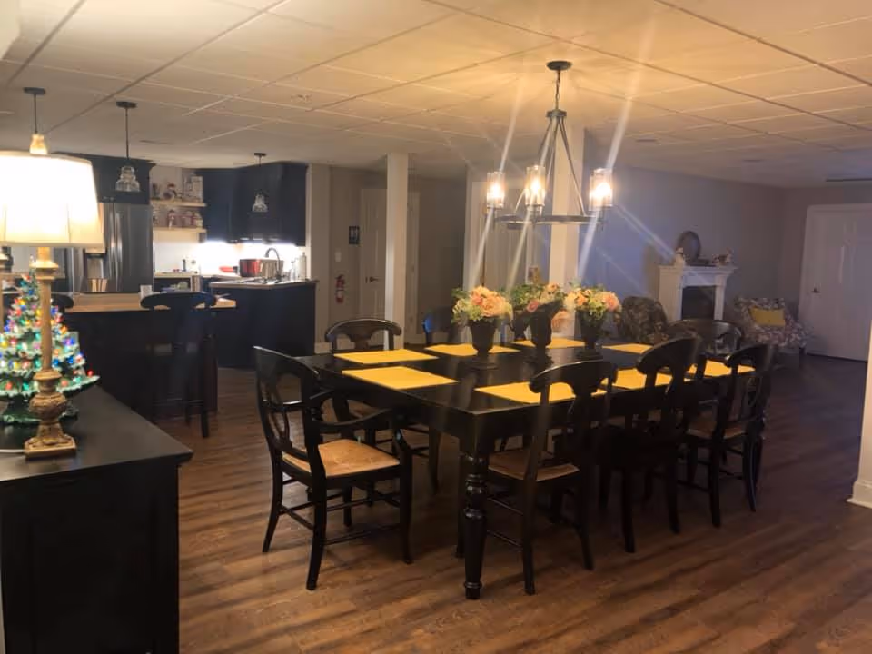 Interior view of a dining area with a large black dining table set with yellow placemats and floral centerpieces. The room has wooden flooring and is softly lit by a chandelier above the table. In the background, there is a kitchen area with black cabinets and a countertop with bar stools. A small decorated Christmas tree and a lamp are visible on a side table to the left.