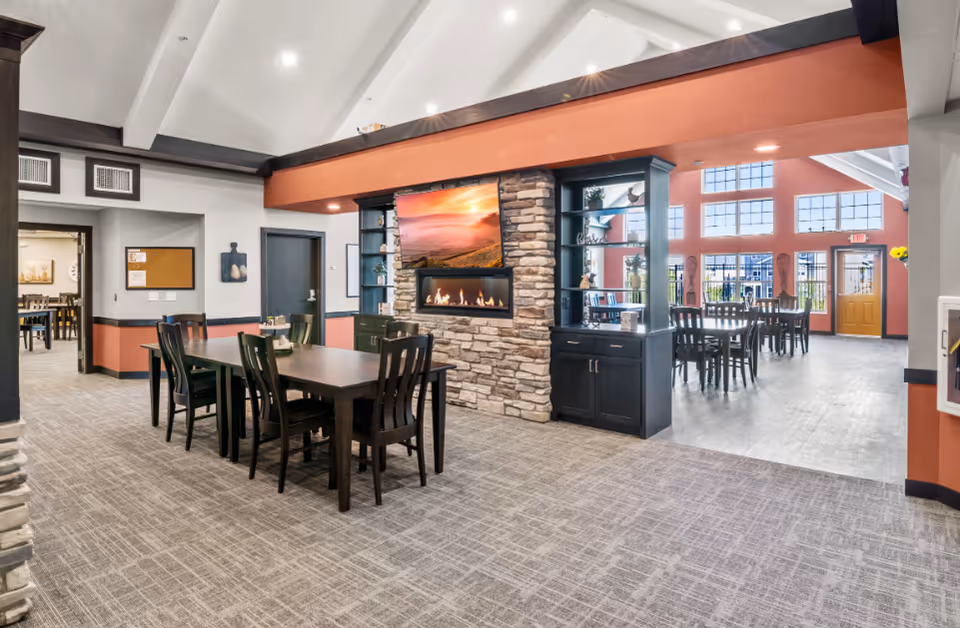A spacious and well-lit dining area in a senior living facility featuring multiple dark wood tables and chairs. The room has a high vaulted ceiling with white beams and a warm color scheme with orange and gray walls. A stone fireplace with a mounted flat-screen TV displaying a sunset scene is centrally located, flanked by built-in dark wood shelving units. Large windows in the background allow natural light to fill the space, and an exit door is visible at the far end. The carpeted floor adds to the cozy ambiance.