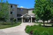 Exterior view of a multi-story senior living facility building with a covered entrance, surrounded by green trees, shrubs, and a paved walkway leading to the entrance under a partly cloudy sky.