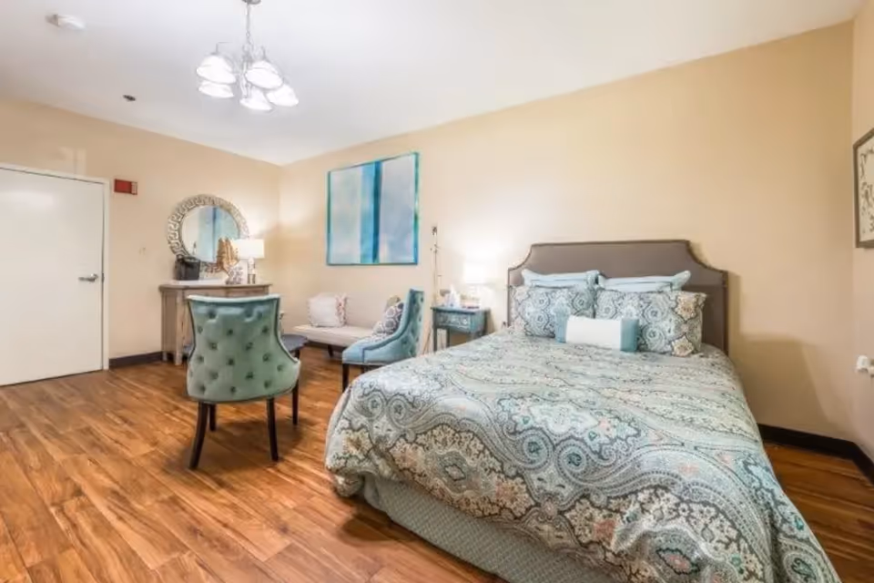 A cozy bedroom with a large bed covered in a patterned bedspread in shades of blue and beige. The room has wooden flooring, a small sitting area with two upholstered chairs and a loveseat, a wooden dresser with a round mirror above it, and soft lighting from table lamps and a ceiling light fixture. The walls are painted a light beige color and there is a piece of abstract artwork hanging above the loveseat.