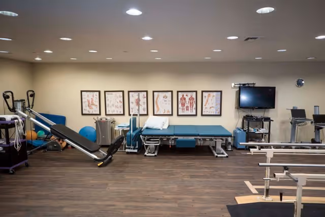 A physical therapy room with exercise equipment including a treatment table, resistance bands, exercise balls, and parallel bars. Anatomical charts are displayed on the wall behind the treatment table, and a television is mounted on the wall to the right.