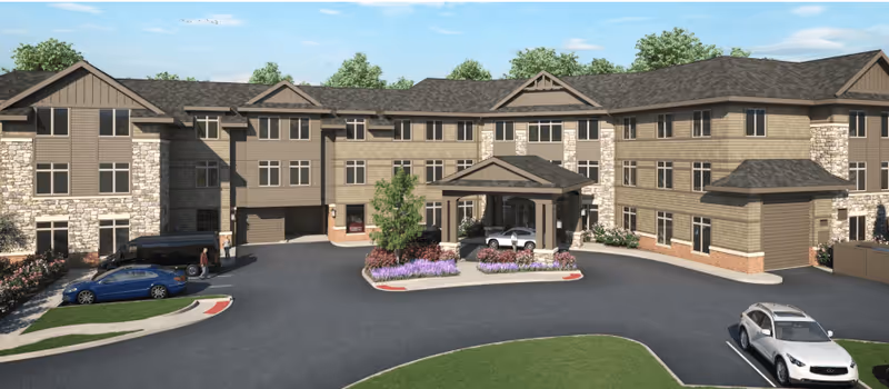 Exterior view of The Kensington Reston senior living facility showing a three-story building with stone and wood siding, multiple windows, a covered entrance with a driveway, landscaped flower beds, and several parked cars.