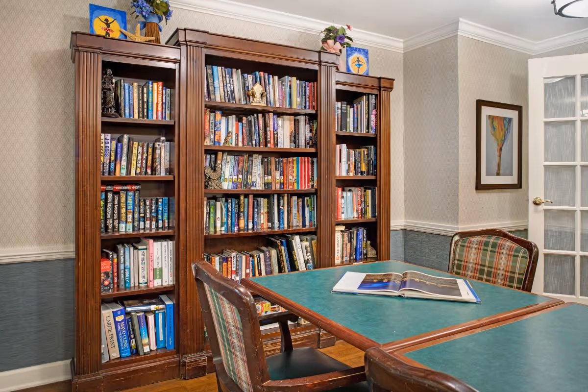 A cozy reading room with tall wooden bookshelves filled with books, a green-topped table and plaid chairs.