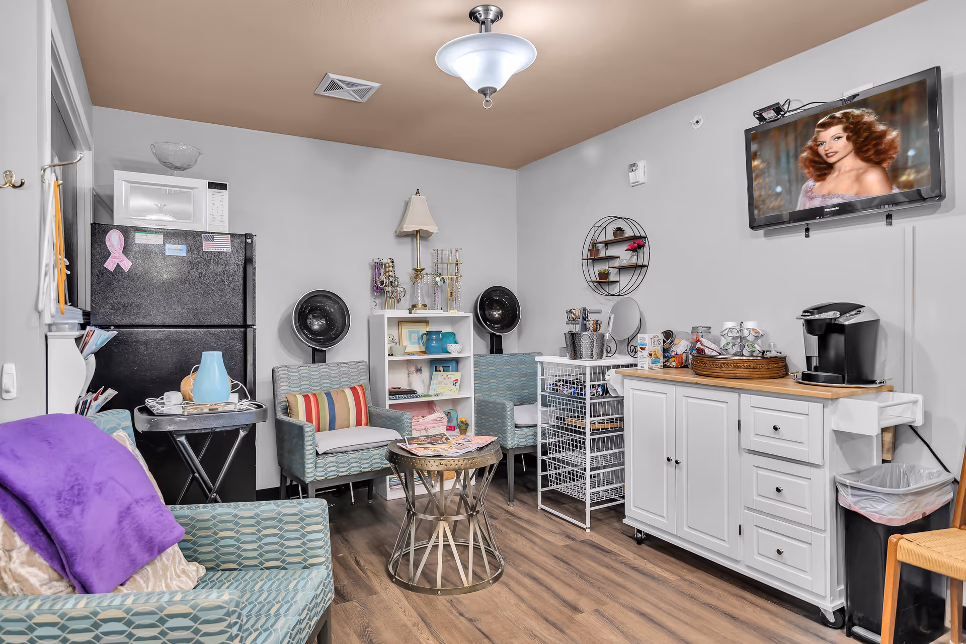 Cozy common room with armchairs, a wall-mounted TV, a refrigerator and coffee station, shelving, and hooded salon dryers.