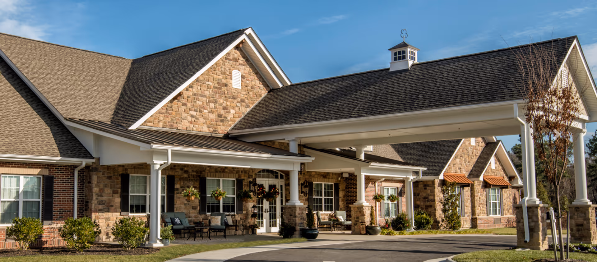 Exterior view of Bickford of Chesterfield facility showing a large covered entrance with stone and brick facade, multiple windows, and outdoor seating areas with chairs and potted plants under a clear blue sky.