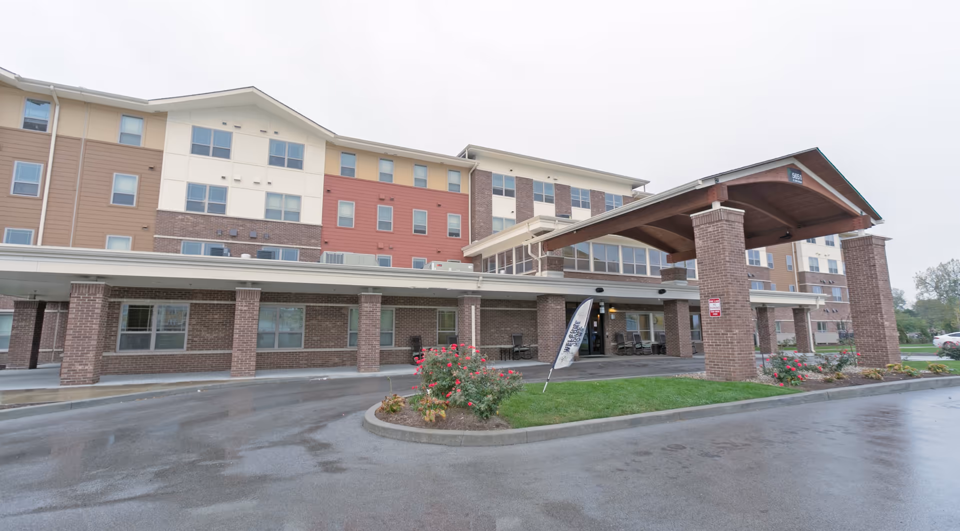 Exterior view of a multi-story senior living facility named Oasis At 30th, featuring a covered entrance with brick pillars, a driveway, and landscaped areas with flowers and grass.