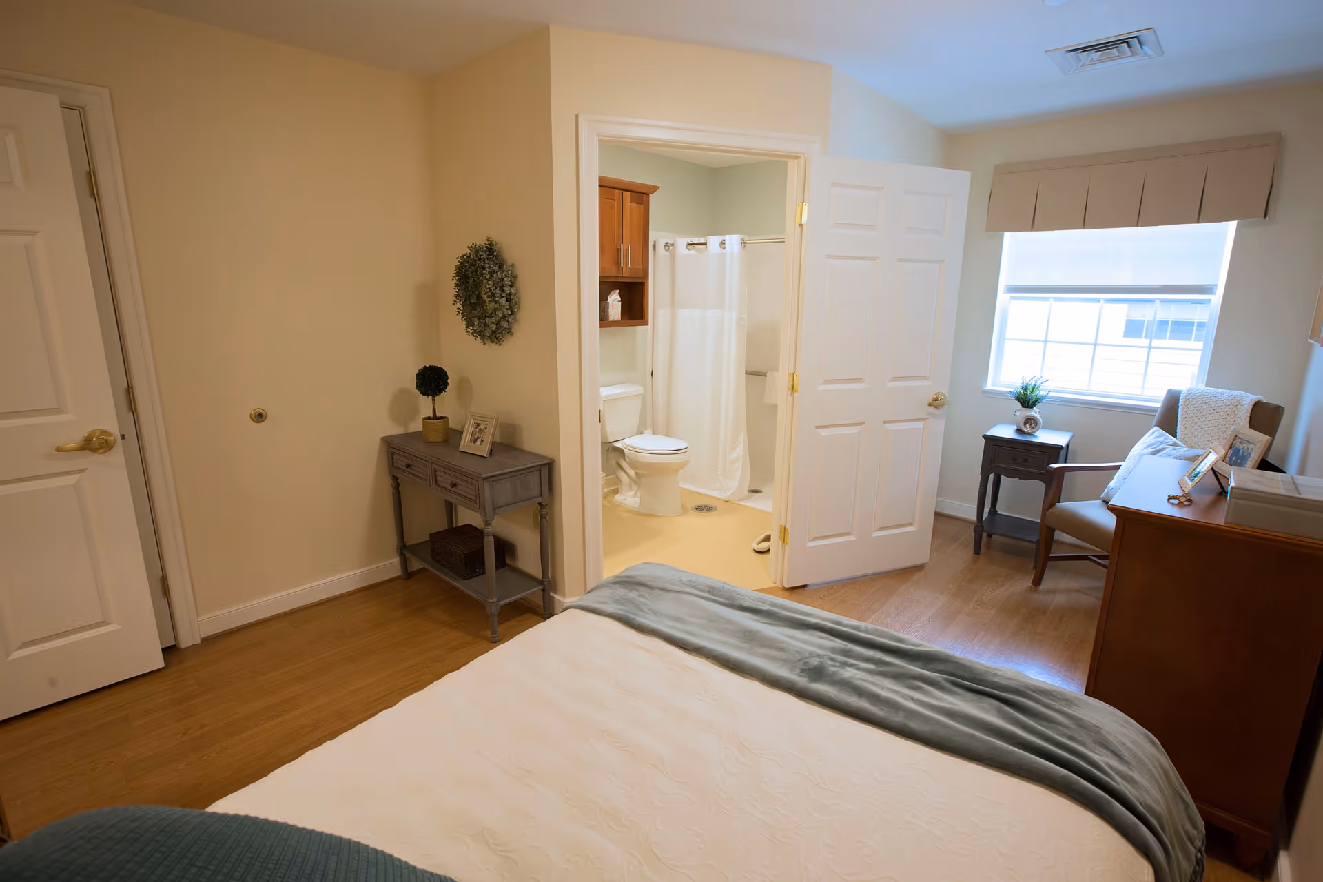 A cozy bedroom in a senior living facility featuring a bed with a white quilt and gray blanket, a small gray console table with decorative items, a wooden dresser, an armchair with a pillow and blanket, and a window with a beige valance. The bedroom has an open door leading to a bathroom with a toilet, shower with white curtain, and wooden cabinet.