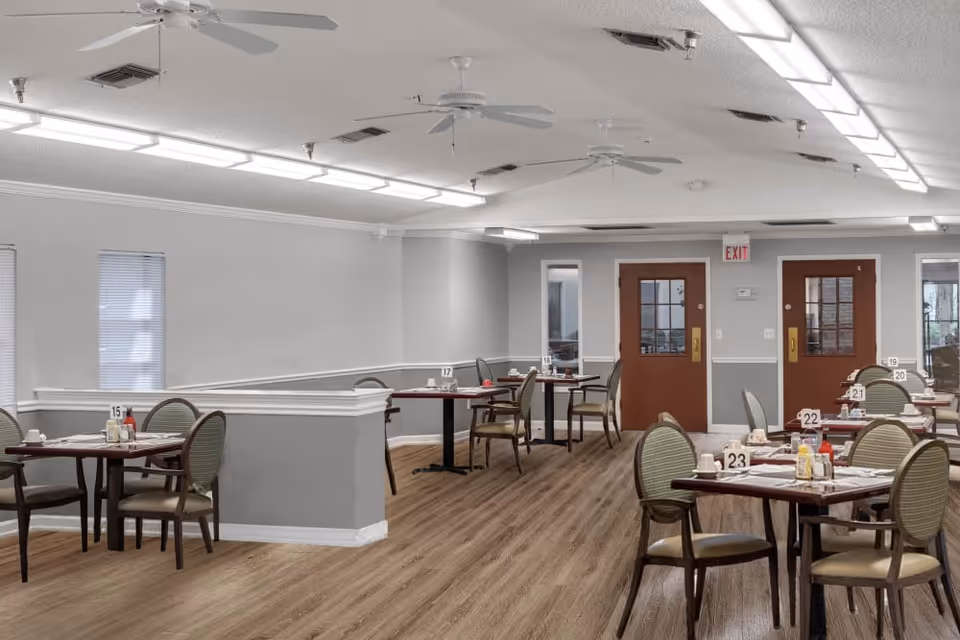 A dining room in a senior living facility with multiple tables and chairs arranged neatly. Each table has place settings, condiments, and table number signs. The room has wood flooring, gray and white walls, ceiling fans, and fluorescent lighting. Two brown doors and windows are visible at the far end of the room.