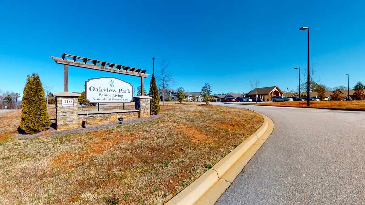 Outdoor view of the entrance to Oakview Park Senior Living facility, showing a large sign with the facility name and description 'Assisted and Memory Care' next to a curved driveway and landscaped area with small trees and bushes under a clear blue sky.