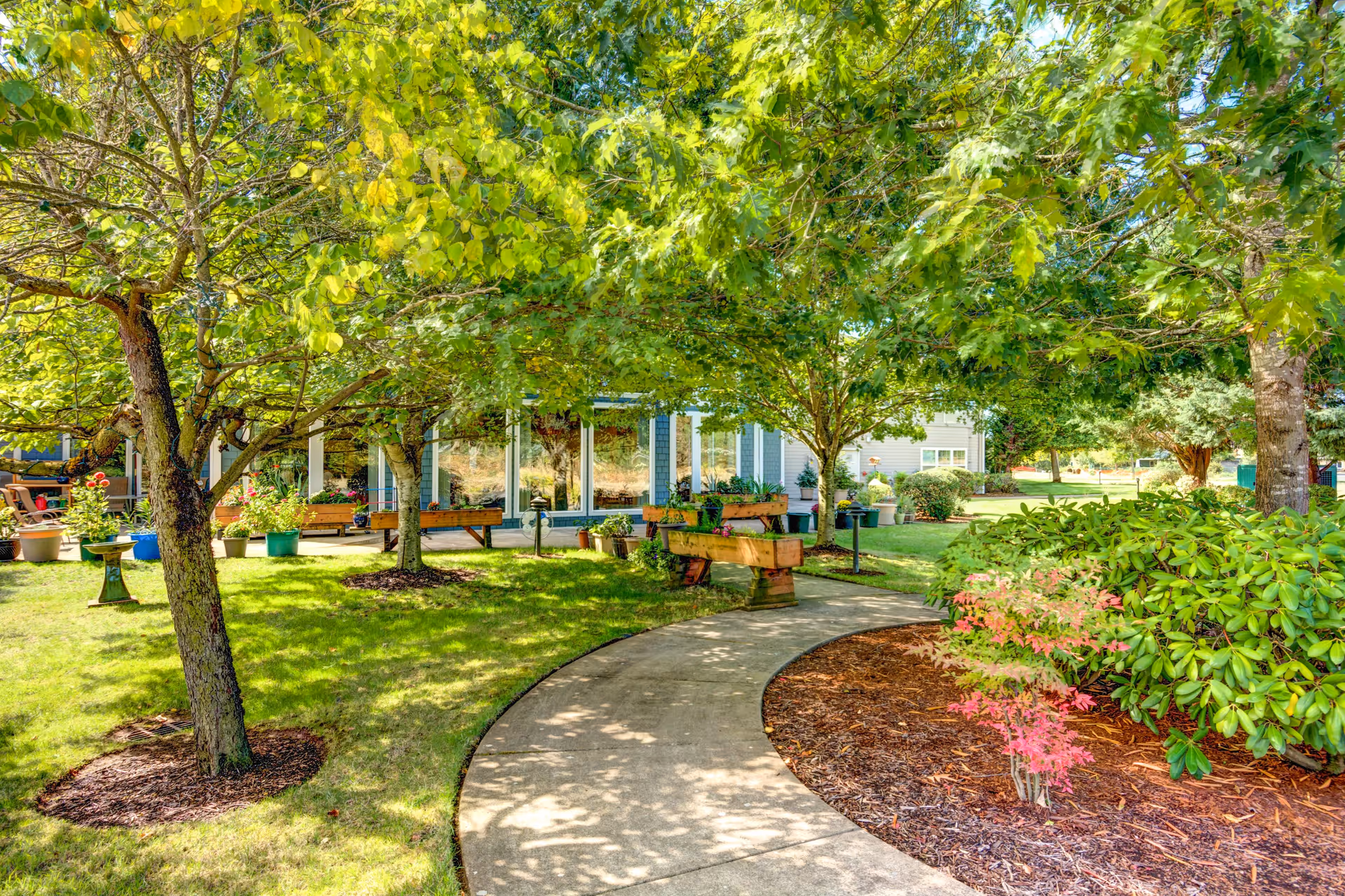 A sunny outdoor garden area at Lone Oak Assisted Living featuring a curved concrete pathway surrounded by green grass, trees, and various potted plants. Wooden benches and planters are placed along the path, with a building visible in the background through the trees.