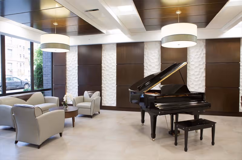 A bright and modern lounge area featuring a black grand piano with a matching bench, surrounded by light gray armchairs and a small round wooden table with a flower vase. The room has large windows letting in natural light, textured white and dark wood panel walls, and two large circular pendant lights hanging from the ceiling.