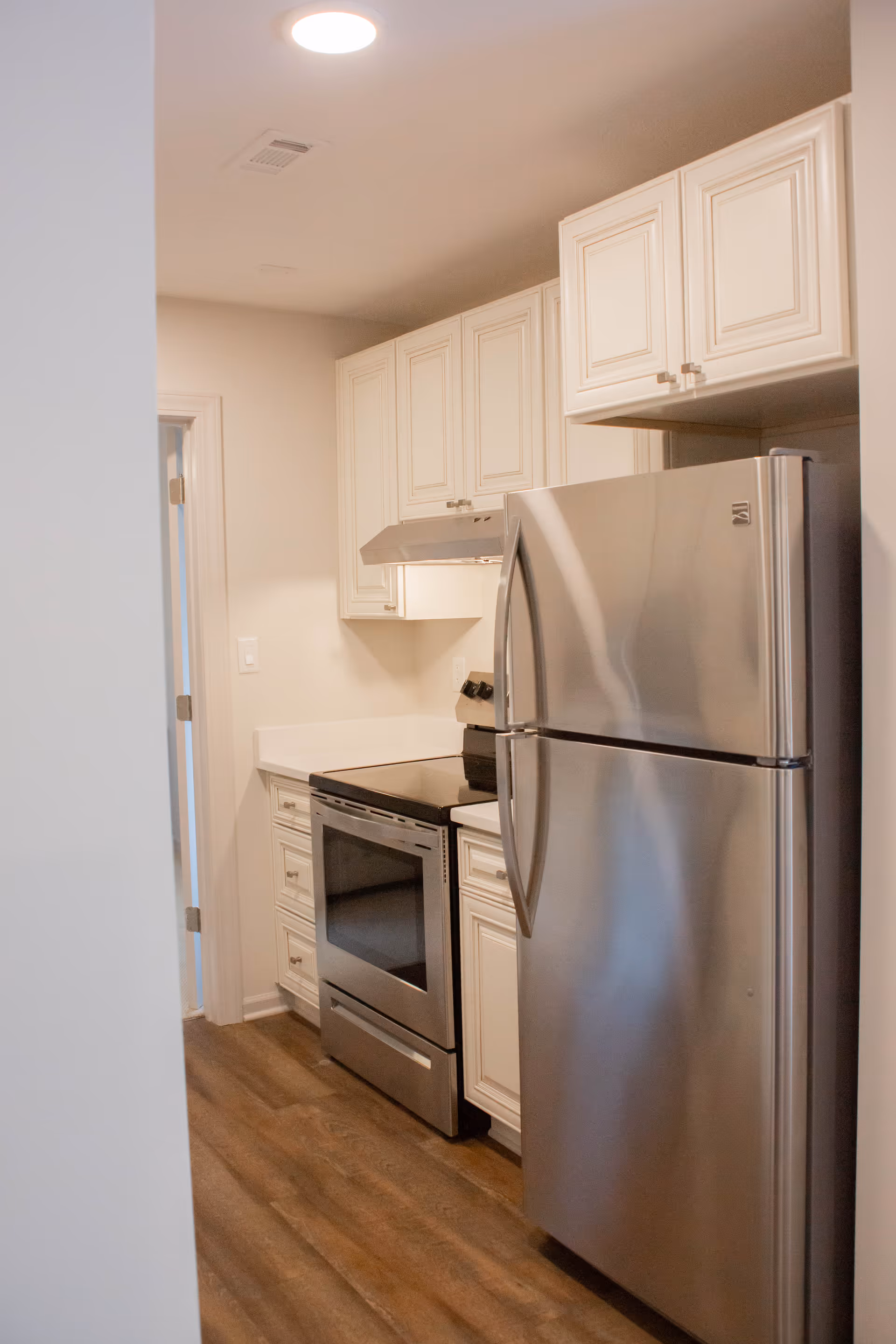 A modern kitchen with white cabinetry, a stainless steel refrigerator, an electric stove with oven, and a range hood. The floor is wood, and the walls are painted light beige.