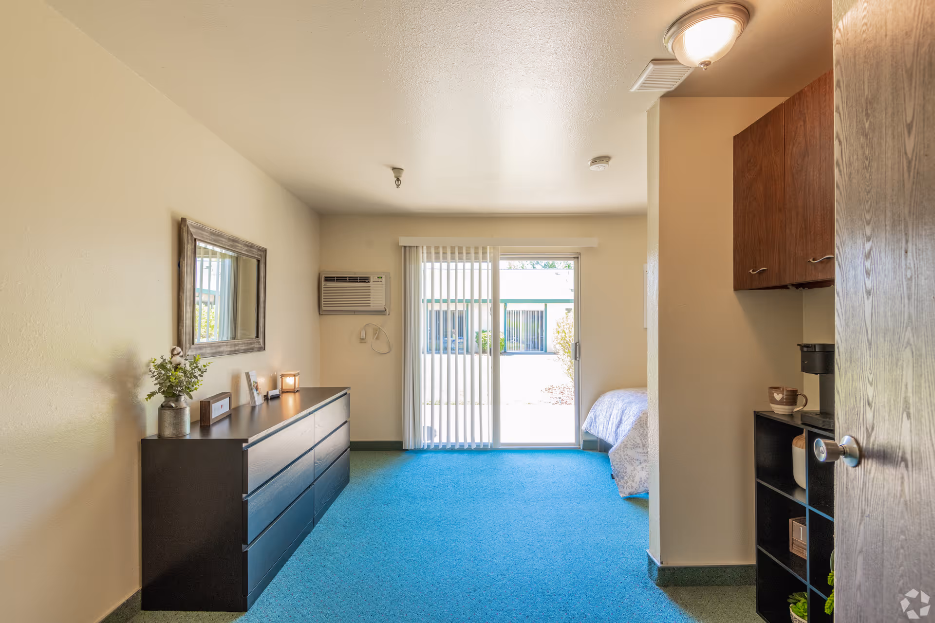 Studio apartment interior with a dresser and mirror, sliding glass door, and a partially visible bed.