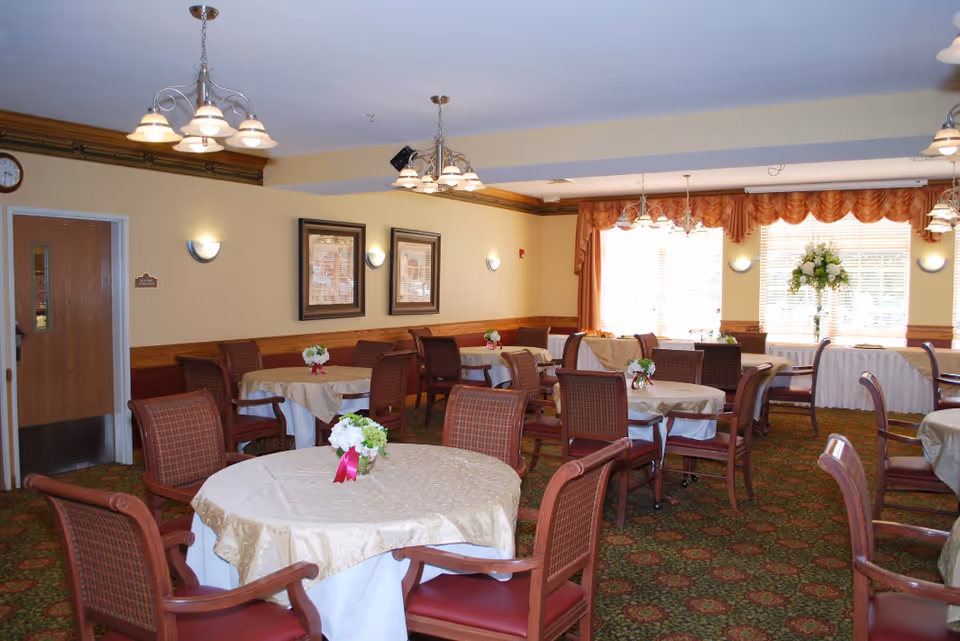 A dining room with multiple round tables covered with beige tablecloths and small floral centerpieces. The room has wooden chairs with red cushions, patterned carpet, and large windows with orange curtains letting in natural light. There are chandeliers and wall sconces providing lighting, and a door on the left side of the room.