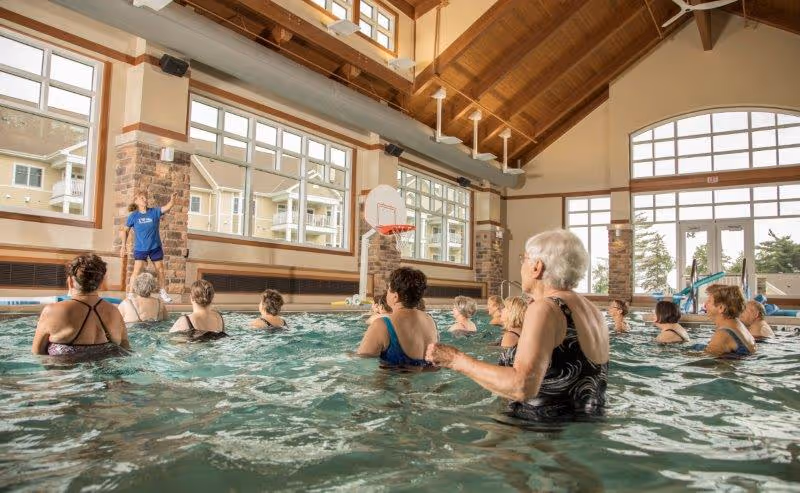 A group of elderly people participating in a water exercise class in an indoor swimming pool. An instructor stands on the pool deck demonstrating exercises. The pool area has large windows, high ceilings with exposed beams, and a basketball hoop mounted on the wall.