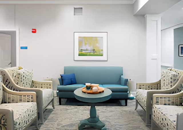 A cozy living room area with a teal sofa adorned with two blue pillows, flanked by two patterned armchairs. A round teal coffee table sits in front of the sofa with a wooden tray holding decorative items. A framed abstract painting hangs on the white wall above the sofa.