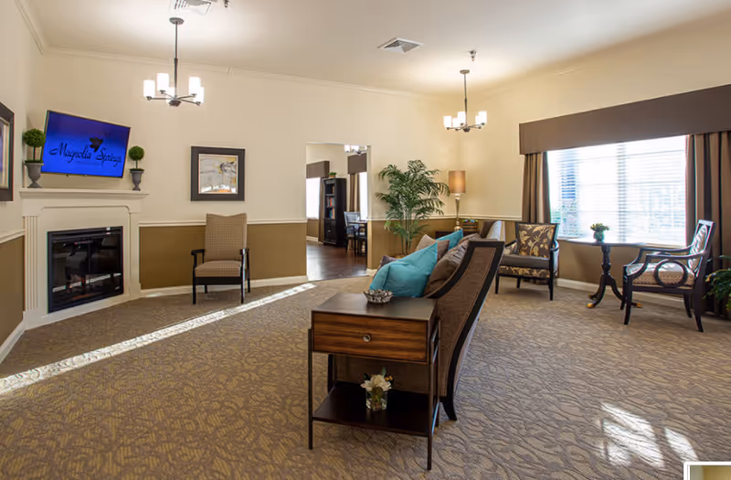 Sunlit communal lounge with sofas, chairs, side tables, a fireplace, and a wall-mounted TV showing 'Magnolia Springs'.