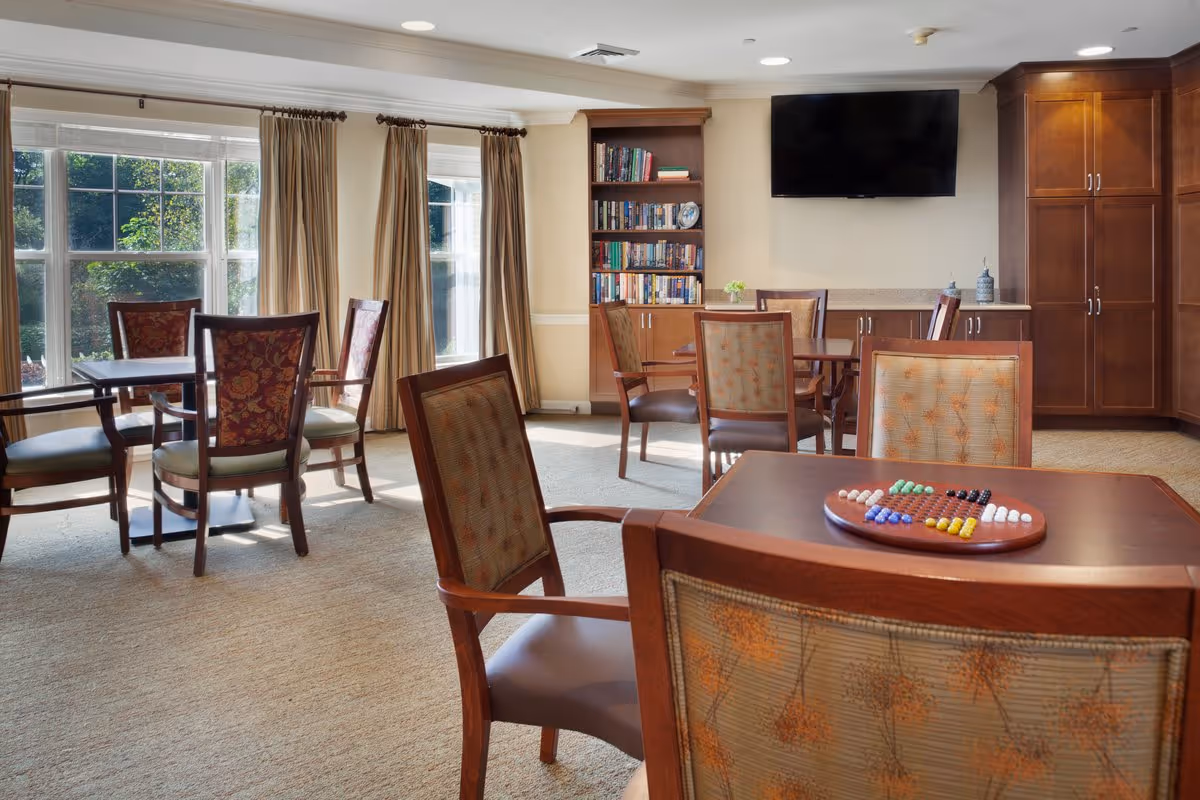 A bright and cozy common area in a senior living facility with several wooden tables and chairs arranged around the room. A large window with beige curtains lets in natural light. There is a bookshelf filled with books and a flat-screen TV mounted on the wall above a cabinet. One table in the foreground has a Chinese checkers game set up.