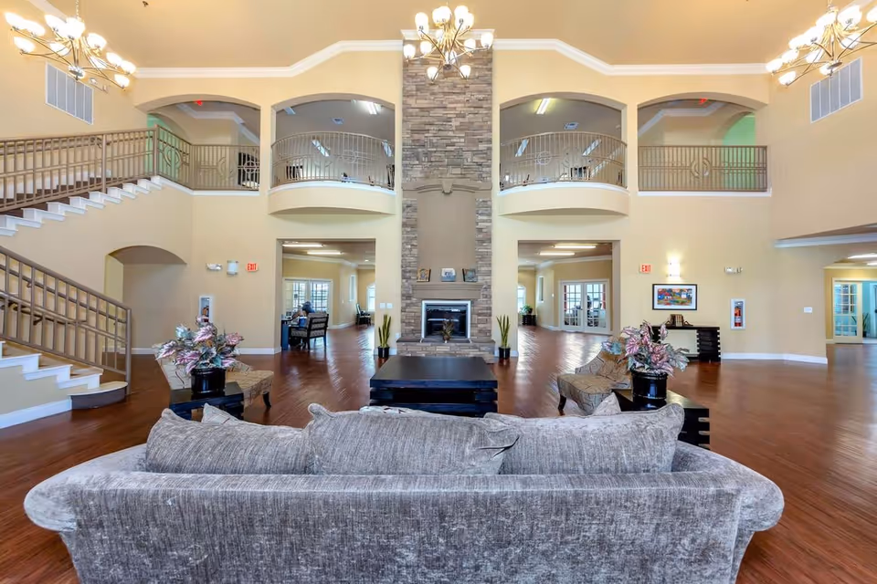 Spacious senior living facility common area with a large gray sofa in the foreground, two armchairs with side tables and plants, a central stone fireplace, and a high ceiling with chandeliers. There is a staircase on the left and upper balcony railings overlooking the area.