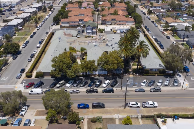 Aerial view of a residential-style building complex with a large parking lot filled with cars in front. The building is surrounded by trees and palm trees, located at the intersection of two streets in an urban neighborhood.