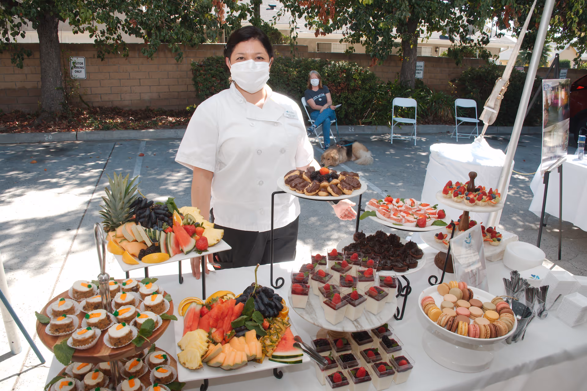 A woman wearing a white chef's coat and a face mask stands behind a table filled with an assortment of desserts and fresh fruit platters outdoors. The table displays mini carrot cakes, macarons, chocolate desserts, fruit cups with berries, and various fresh fruit slices including pineapple, watermelon, cantaloupe, and grapes. In the background, a woman wearing a mask sits on a chair with a dog lying on the ground beside her. Trees and a brick wall are visible behind them.