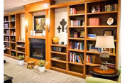 Cozy interior of a senior living facility featuring wooden bookshelves filled with books and decorative items, a fireplace with a painting above it, a small wooden stool, potted plants on the floor, and a table lamp on a round wooden side table.