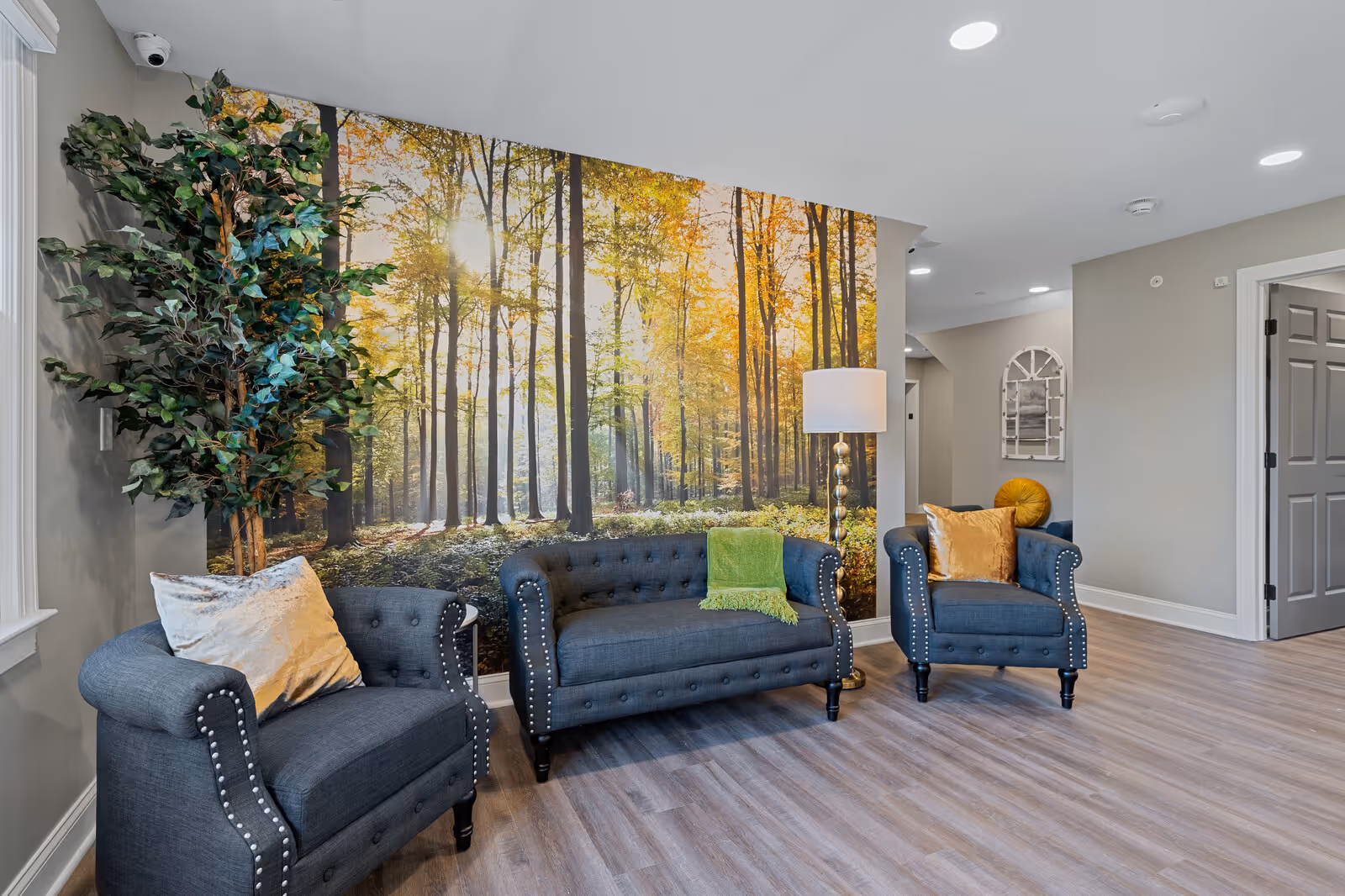A cozy seating area in a senior living facility featuring three dark gray upholstered chairs with studded detailing and colorful throw pillows. Behind the chairs is a large wall mural depicting a sunlit forest with tall trees and autumn foliage. A tall floor lamp with a white shade stands next to the middle chair, and a large artificial plant is placed in the corner near a window. The room has light wood flooring and neutral-colored walls with recessed lighting in the ceiling.