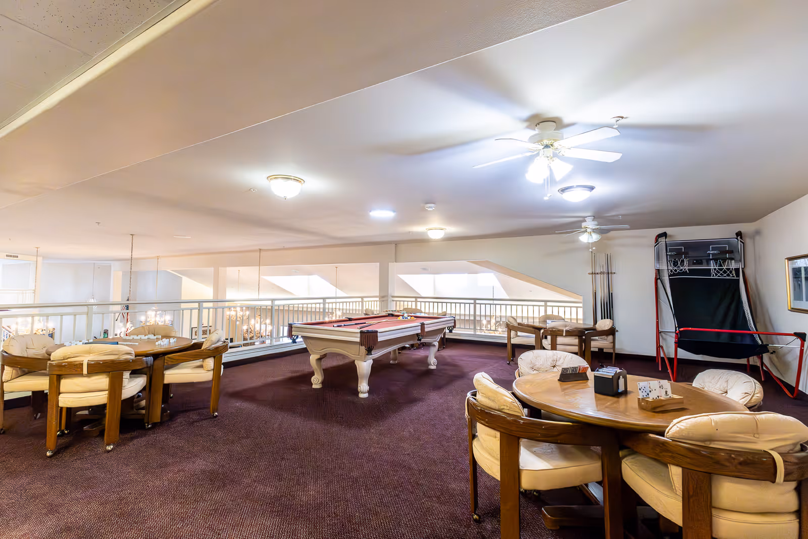 A carpeted communal recreation room with a pool table, card tables and an arcade basketball game.