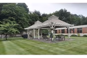 Covered wooden gazebo with tables and chairs on a manicured lawn in front of a brick building and trees.