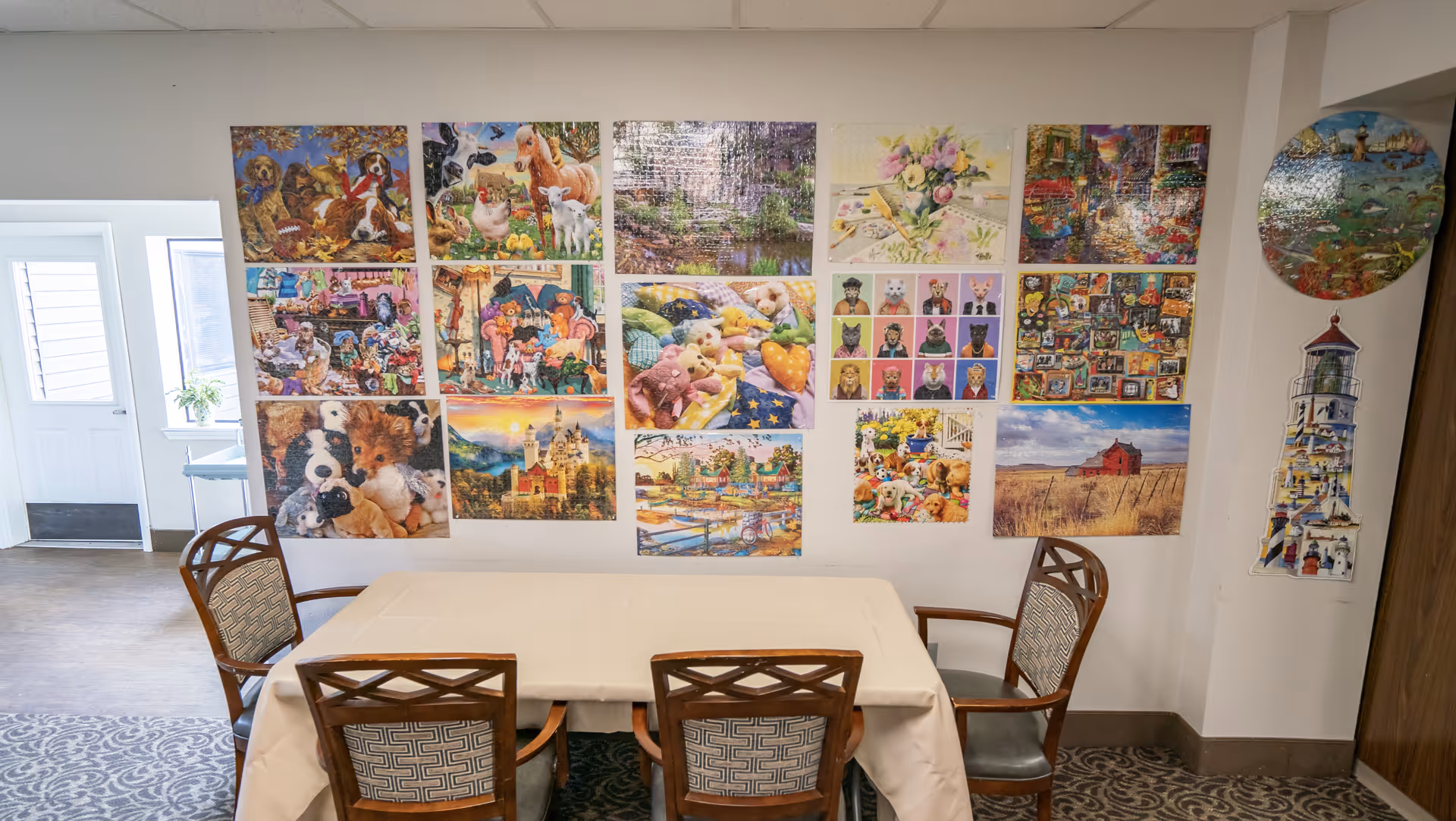 A small dining area with a rectangular table covered with a beige tablecloth and four wooden chairs with patterned cushions. The wall behind the table is decorated with multiple colorful puzzle images featuring animals, landscapes, and various scenes. To the left, there is a door with a window and a small table with a plant on it. The floor has a patterned carpet.
