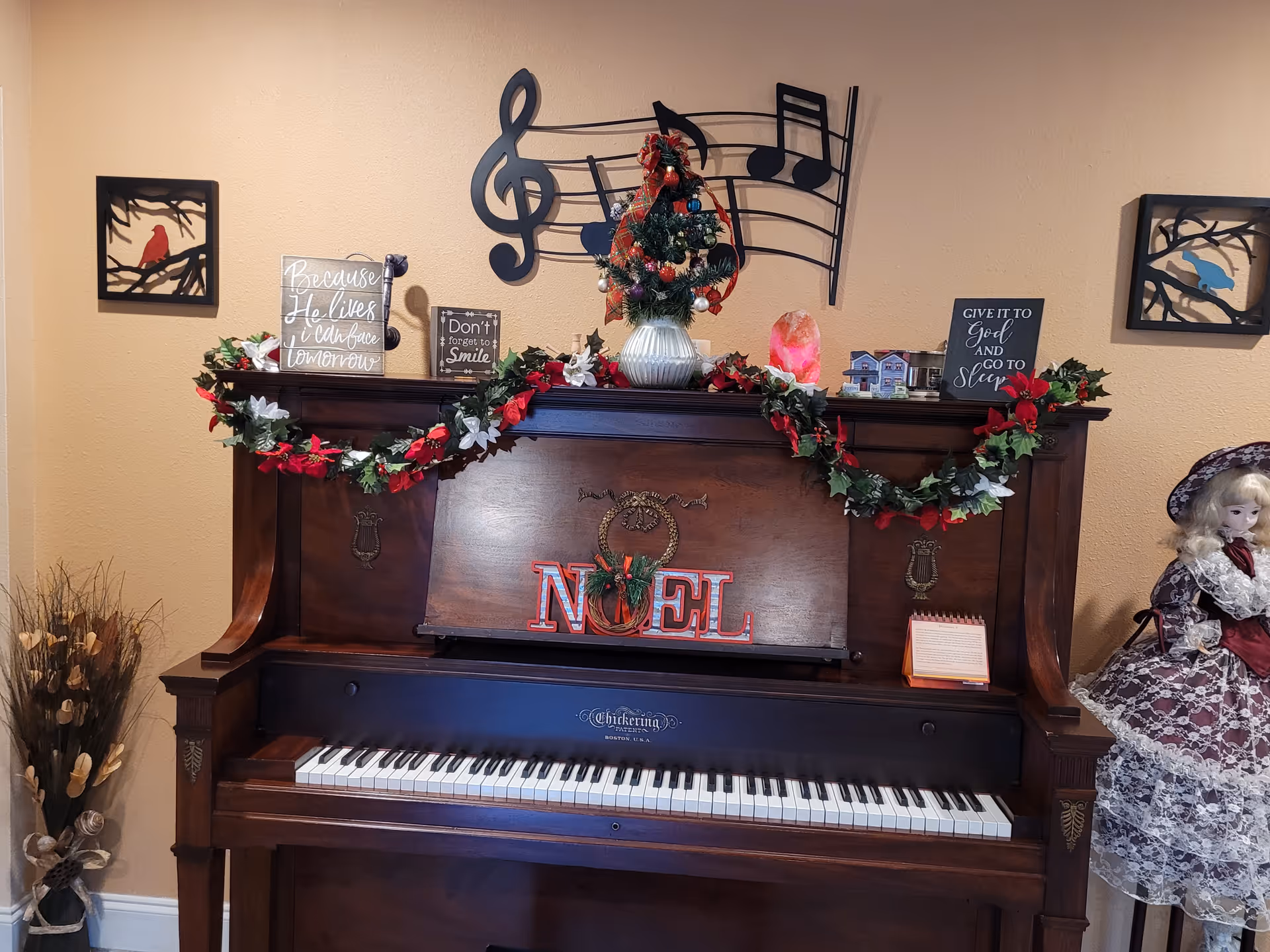 A decorated upright piano with a garland of red and white flowers draped across the top. On the piano are various decorative items including a small Christmas tree, a sign that says 'NOEL', and several inspirational quote plaques. Above the piano is a large black metal wall decoration shaped like musical notes on a staff. To the right of the piano is a porcelain doll dressed in a Victorian-style outfit. On the left side, there is a vase with dried plants. The wall behind is painted beige.
