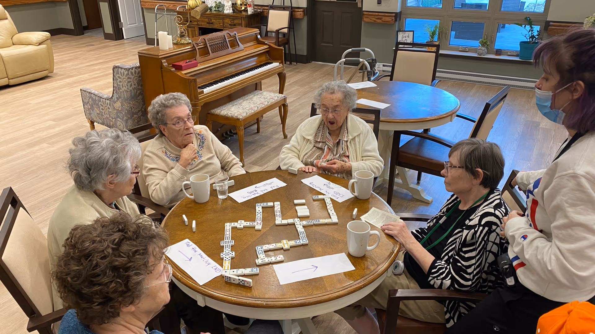 A group of elderly women sitting around a round wooden table playing dominoes in a well-lit common area with wooden floors. A caregiver wearing a mask stands nearby. In the background, there is a piano, chairs, and large windows letting in natural light.