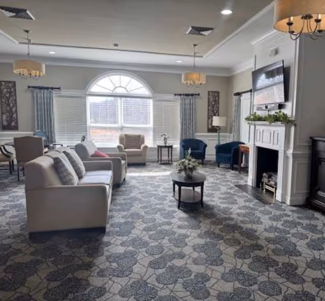 Spacious furnished common living room with sofas and armchairs facing a fireplace and wall-mounted TV beneath a large arched window.