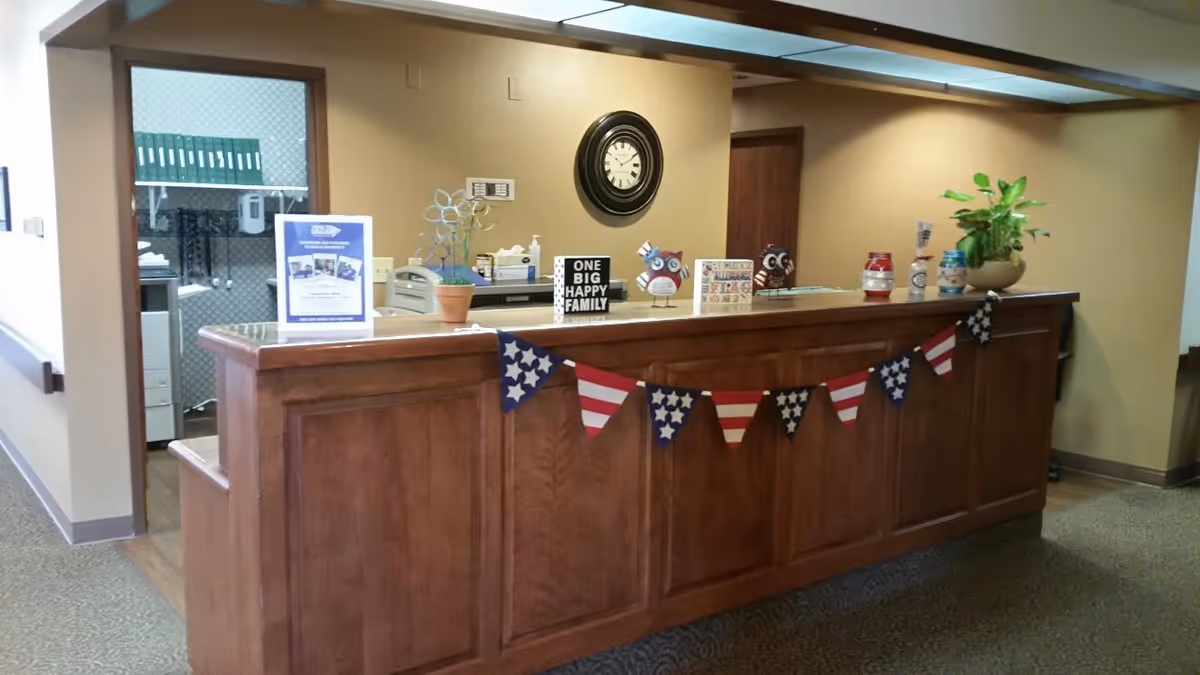 Reception desk area with a wooden counter decorated with a string of American flag-themed pennants. On the counter are various decorative items including a small potted plant, owl figurines, a clock on the wall behind, and a sign that reads 'ONE BIG HAPPY FAMILY'. The background shows an office space with shelves and a printer.