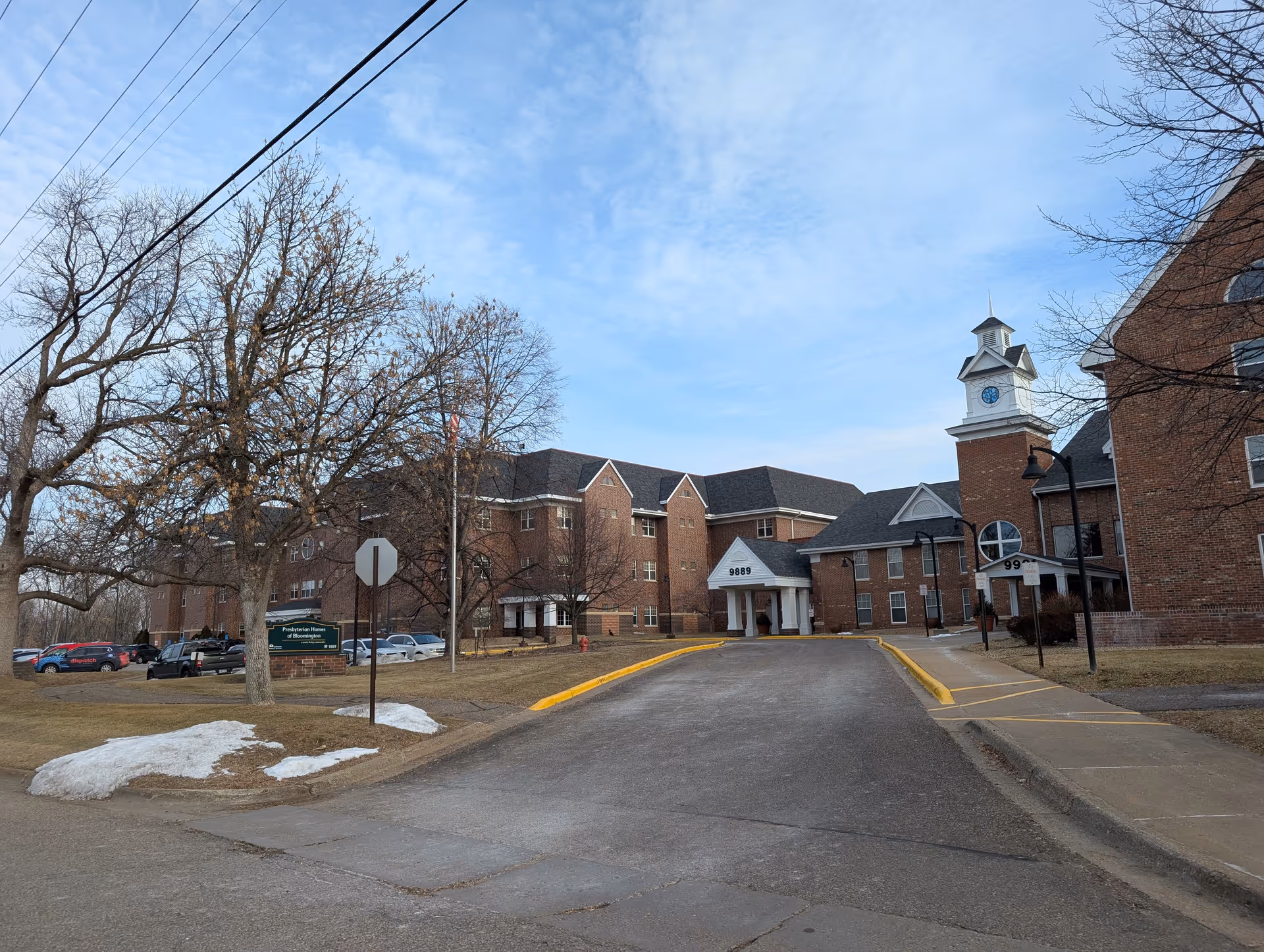 Exterior view of Presbyterian Homes of Bloomington, a large brick building with multiple stories and a clock tower. There is a driveway leading up to the entrance with the number 9889 displayed above it. Leafless trees and patches of snow are visible on the grass surrounding the building under a partly cloudy sky.