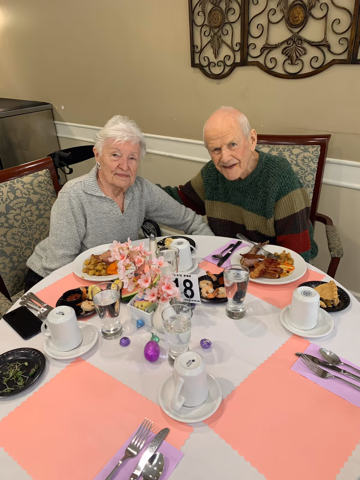 An elderly woman and man sitting together at a round dining table in a senior living facility. The table is set with plates of food, glasses of water, coffee cups, and silverware on pink and white placemats. There is a small vase with pink flowers and some decorative Easter eggs on the table. The background shows a beige wall with decorative metal wall art.