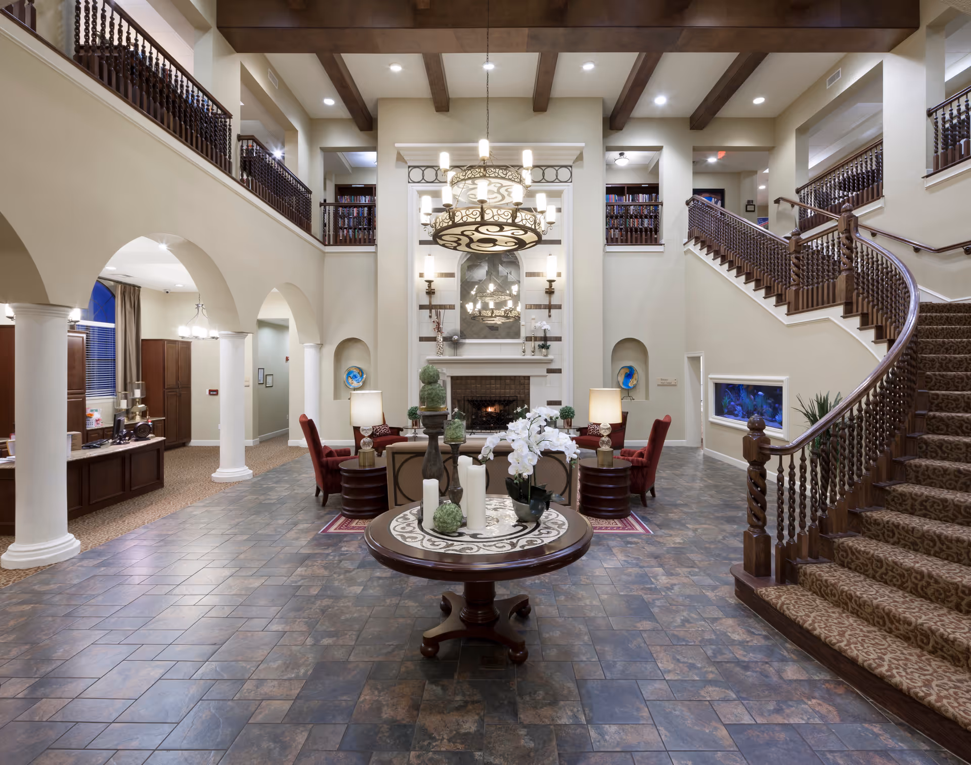 Spacious two-story lobby with a central round table and floral arrangement, chandelier, seating areas, fireplace, and curved staircase.