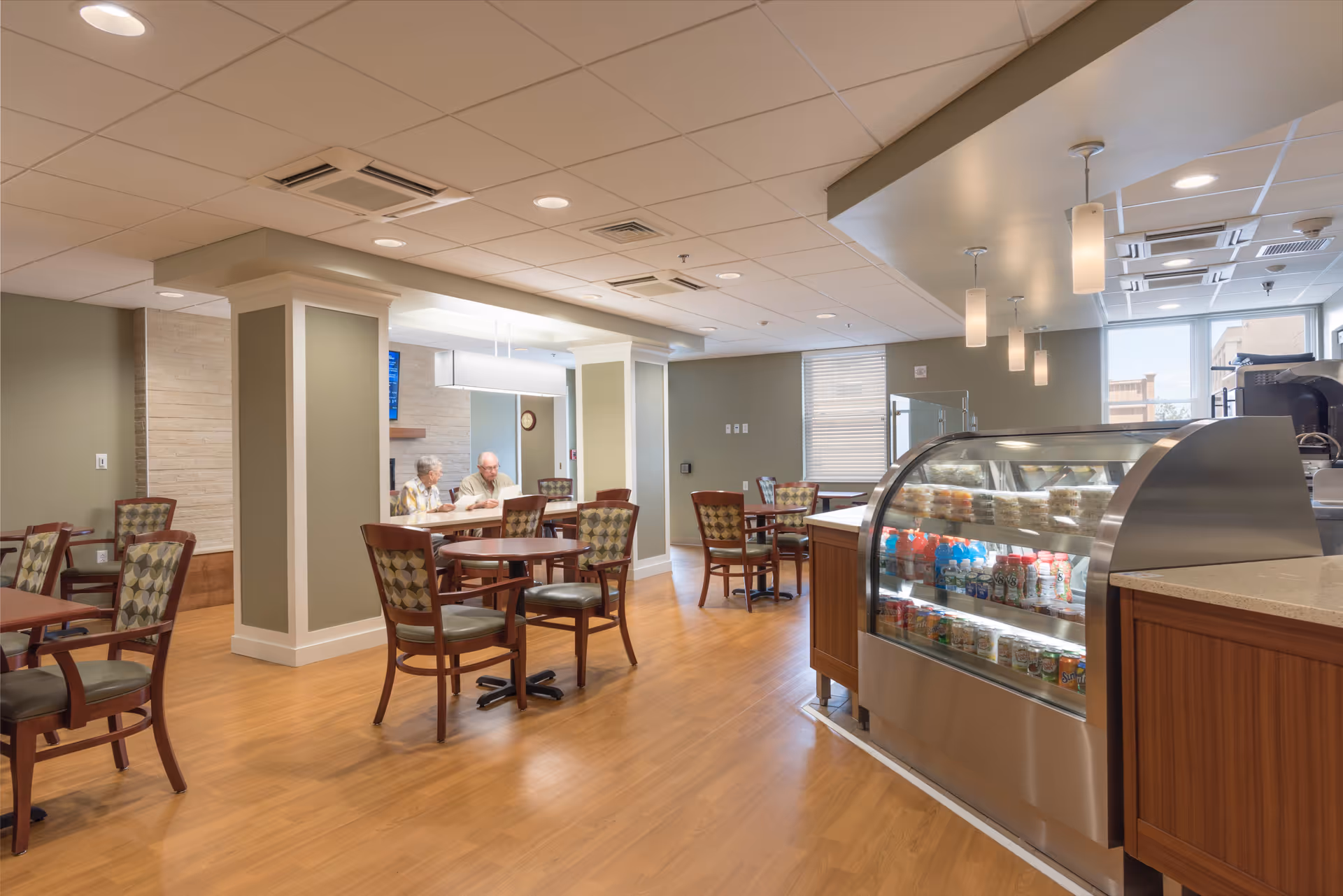 Interior view of a dining area in a senior living facility with several wooden tables and chairs. A refrigerated display case with beverages and snacks is on the right side. Two elderly people are seated at a table in the background, engaged in conversation. The room has wood flooring, soft lighting, and large windows allowing natural light.