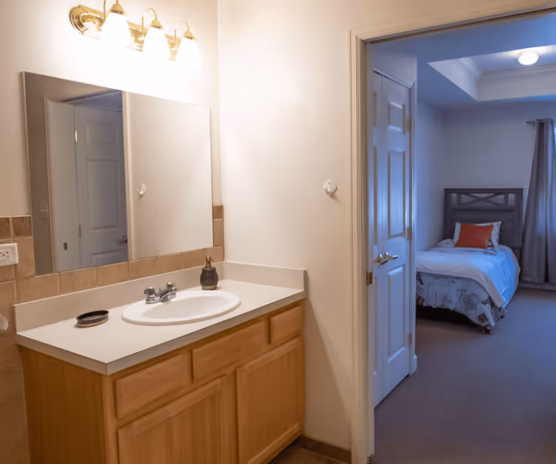 A bathroom vanity with a sink, faucet, soap dispenser, and a large mirror above it. The vanity has wooden cabinets and a white countertop. To the right, an open door leads to a bedroom with a bed, a red pillow, and a window with curtains.