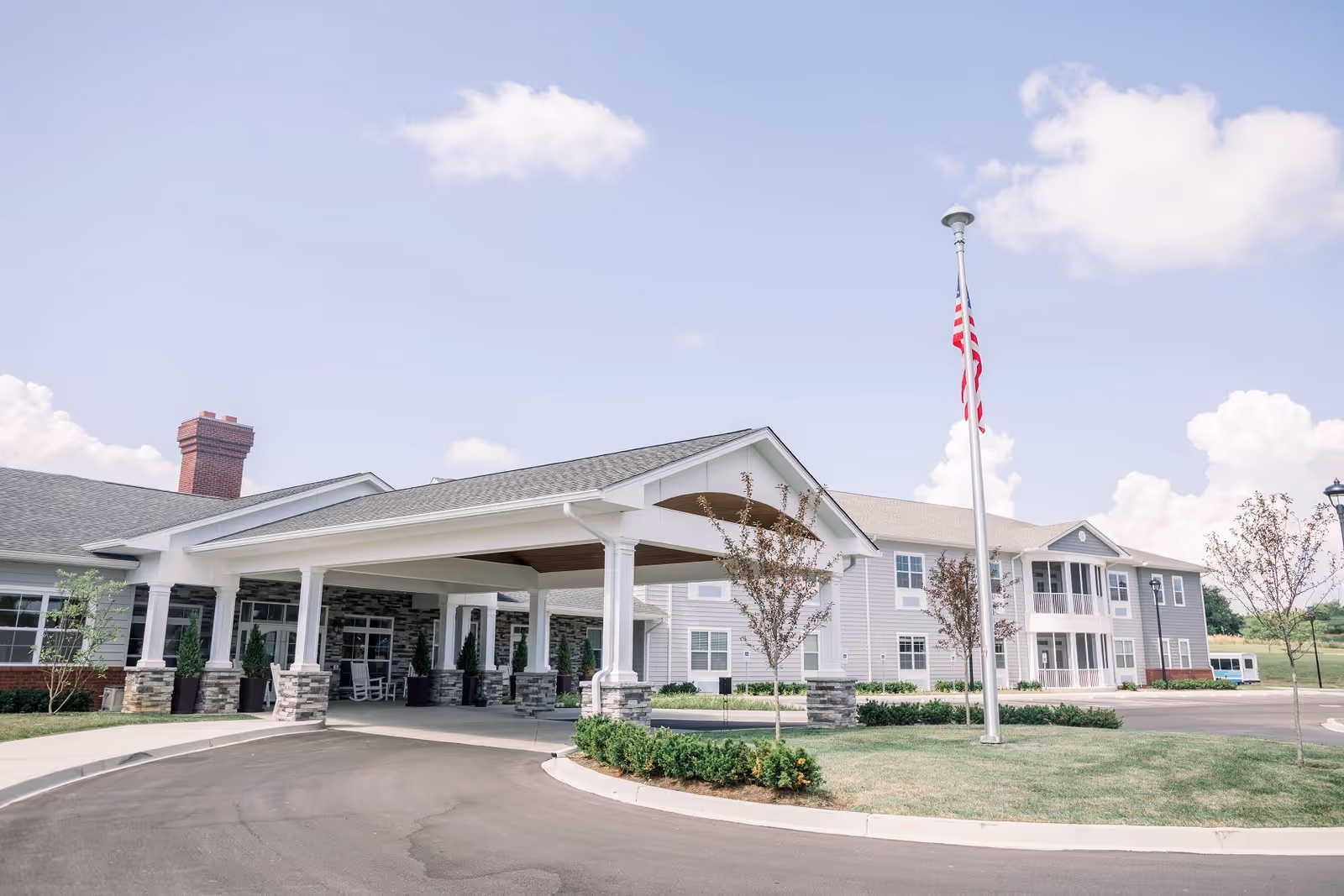 Exterior view of Bluegrass Senior Living facility showing the main entrance with a covered driveway, stone and white pillars, an American flag on a flagpole, and a two-story building with multiple windows under a partly cloudy sky.