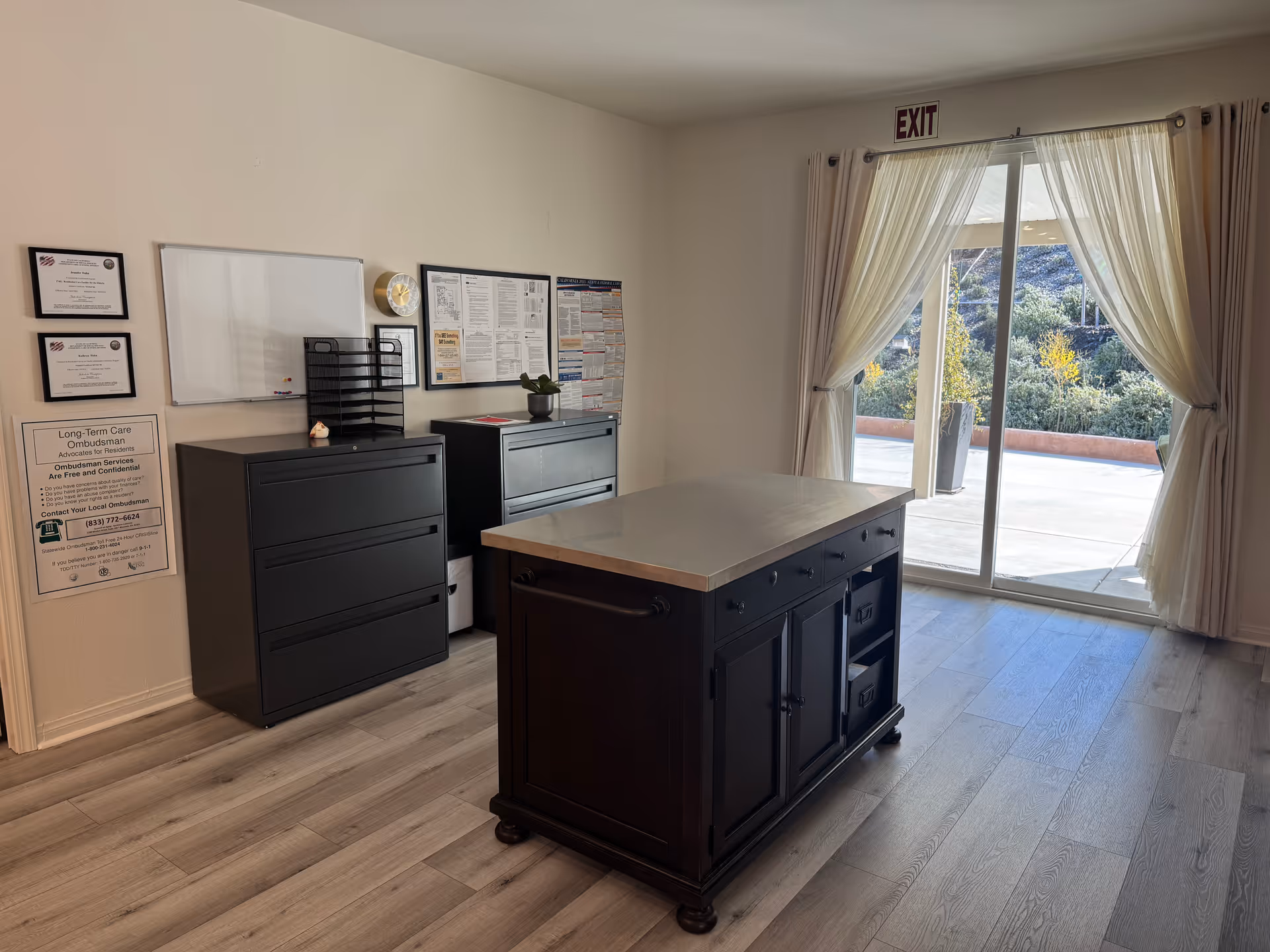 A bright room with light wood flooring featuring a dark wooden island table in the center. Against the wall are two black filing cabinets with various notices, a whiteboard, and a clock above them. A large sliding glass door with sheer curtains opens to an outdoor patio with greenery visible outside. An exit sign is above the door.