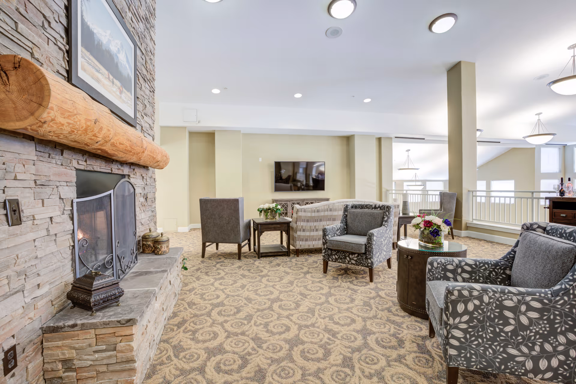A cozy senior living room with patterned carpet, several upholstered armchairs, a stone fireplace with a wooden mantle, a flat-screen TV mounted on the wall, and decorative flowers on tables. The room is well-lit with ceiling lights and natural light from windows.