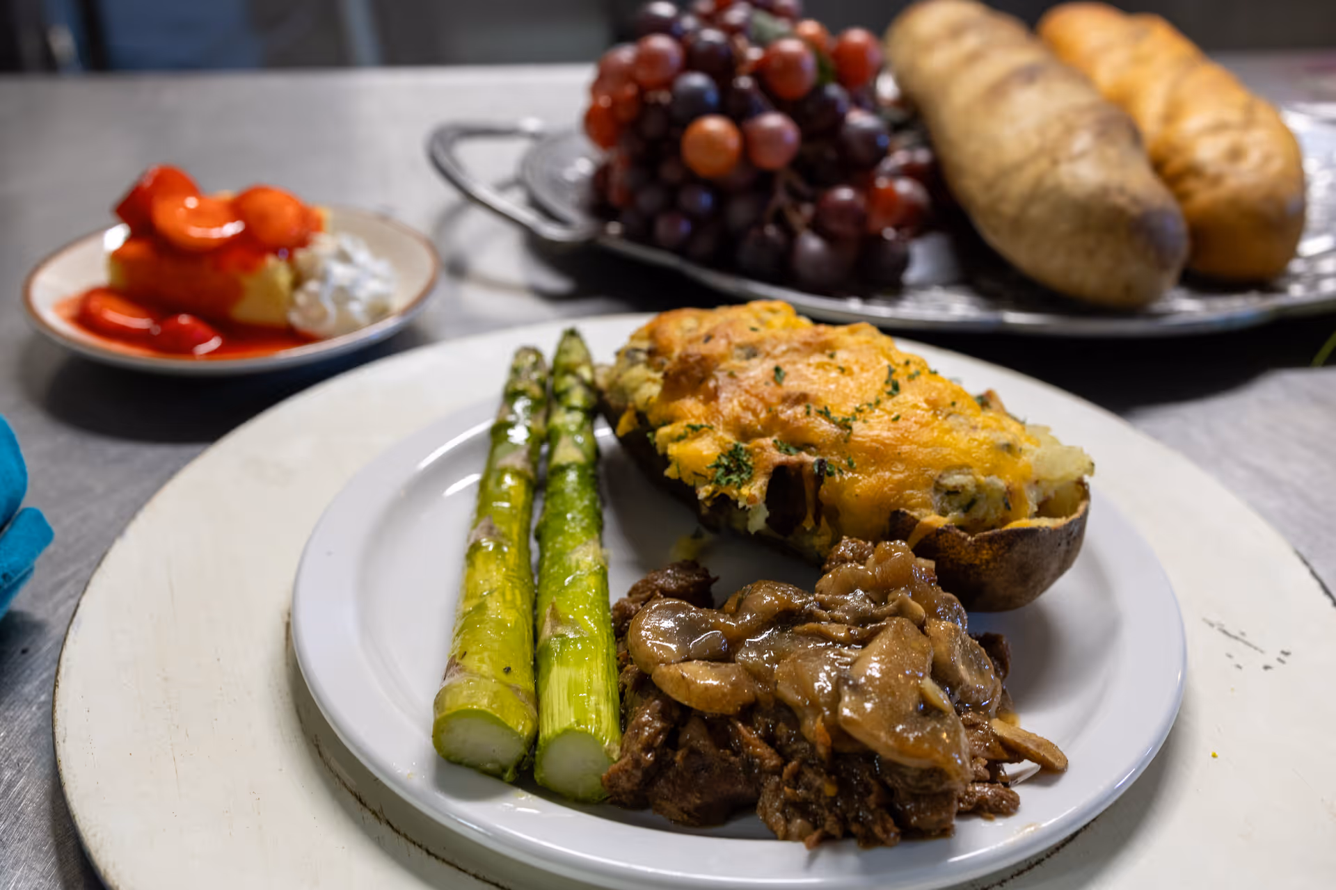 A white plate with two grilled asparagus spears, a twice-baked potato topped with melted cheese and herbs, and a serving of beef with sautéed mushrooms. In the background, there is a plate with red grapes and two whole potatoes, and a small dish with a stuffed pepper and cottage cheese.