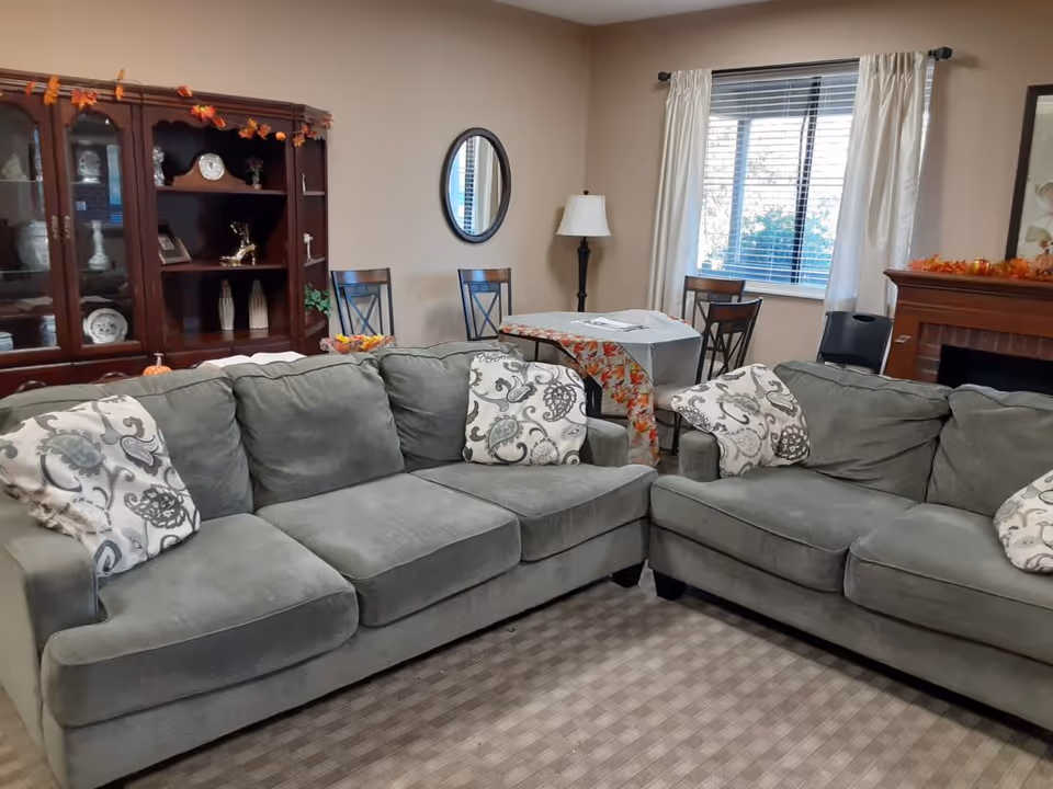 A cozy common living room with two gray sofas and patterned pillows, a small dining table, wooden cabinet, and fireplace.
