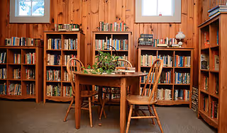 Cozy wood-paneled reading room with bookshelves lining the walls and a round wooden table with chairs and a plant on top.