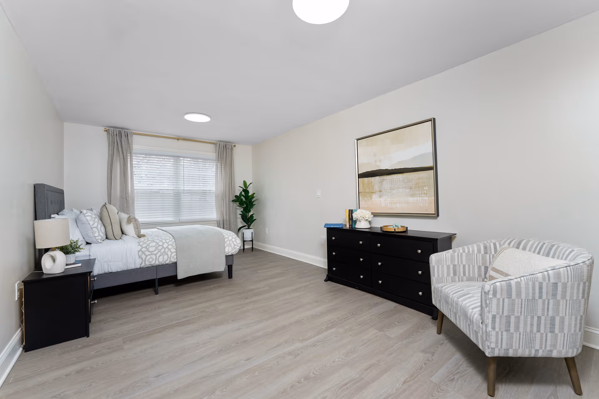 A bright and spacious bedroom with a large window covered by light curtains. The room features a neatly made bed with patterned bedding and multiple pillows, a black nightstand with a lamp and plant, a black dresser with decorative items and books, a modern abstract painting on the wall, and a striped upholstered armchair. The floor is light wood, and the walls are painted white.