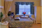 A cozy nursing home room with a single bed covered in a patterned bedspread, a small table with a vase of colorful flowers in front of a window with blue curtains, a microwave on a counter, a beige armchair, and a wooden wardrobe.