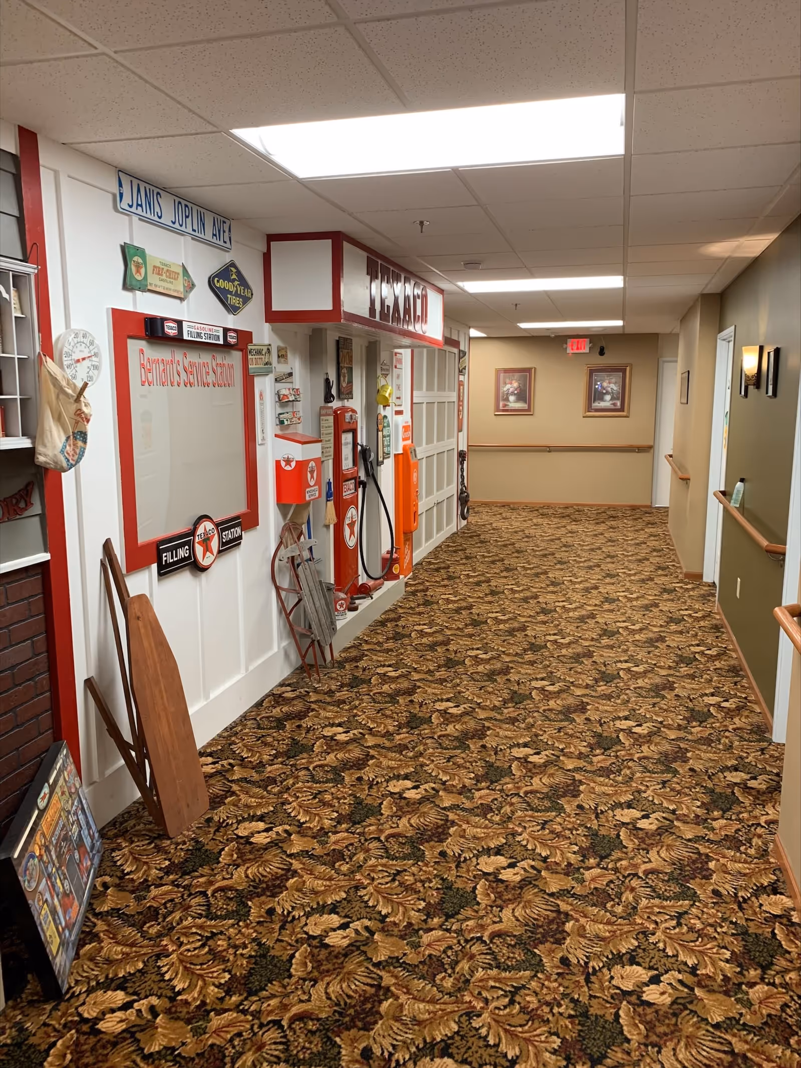 A hallway in a senior care facility decorated with vintage Texaco gas station memorabilia, including signs, a gas pump, and other retro items. The hallway has patterned carpet, beige walls with wooden handrails, and framed pictures at the far end. The ceiling has fluorescent lighting panels.